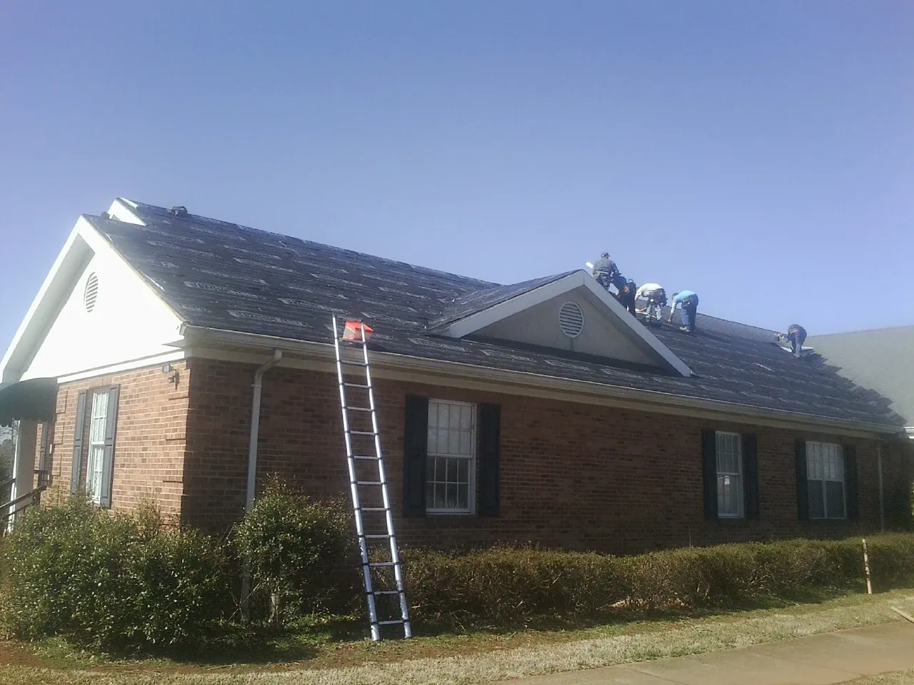 Crew tearing shingles off a brick ranch roof with a ladder set — Metal Roof Repair work by our crew in Wynnewood